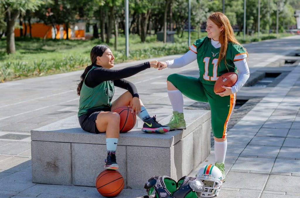 Two female athletes share a fist bump while sitting outdoors, holding sports equipment in Mexico City.